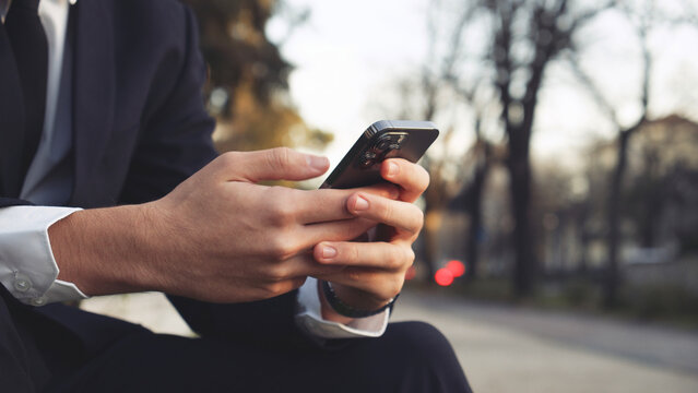 Close up Male hands with smartphone, outdoors. Businessman is typing text messages on mobile phone sitting in the park. Human holds cell phone close up. Man holds mobile phone. Connection concept.