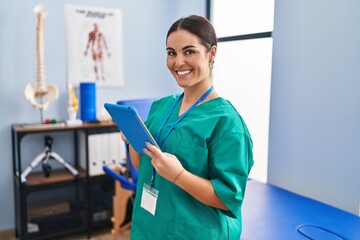 Young beautiful hispanic woman physiotherapist smiling confident using touchpad at rehab clinic
