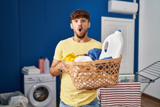 Arab man with beard holding laundry basket and detergent bottle afraid and shocked with surprise and amazed expression, fear and excited face.