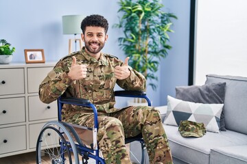 Arab man wearing camouflage army uniform sitting on wheelchair success sign doing positive gesture with hand, thumbs up smiling and happy. cheerful expression and winner gesture.