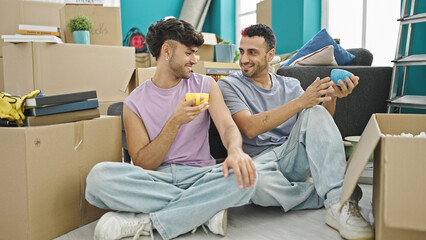 Two men couple drinking coffee sitting on floor speaking at new home
