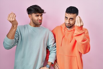 Young hispanic gay couple standing over pink background doing italian gesture with hand and fingers confident expression