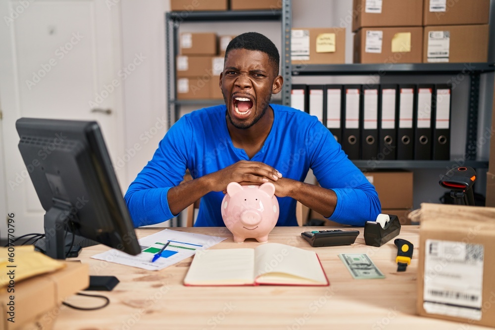 Poster African american man working at small business ecommerce with piggy bank angry and mad screaming frustrated and furious, shouting with anger. rage and aggressive concept.