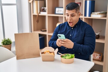 Hispanic young man eating take away food using smartphone celebrating crazy and amazed for success with open eyes screaming excited.