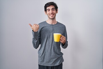 Young hispanic man wearing pajama drinking a cup of coffee pointing to the back behind with hand...