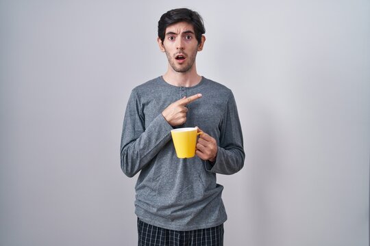 Young hispanic man wearing pajama drinking a cup of coffee surprised pointing with finger to the side, open mouth amazed expression.