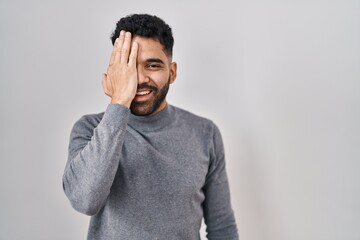 Hispanic man with beard standing over white background covering one eye with hand, confident smile on face and surprise emotion.