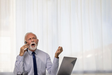 Angry senior businessman shouting, screaming and gesturing during cellphone conversation in front of a computer display in the office