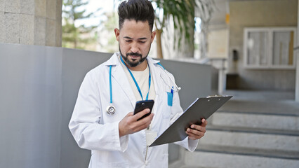 Young latin man doctor using smartphone holding clipboard at hospital