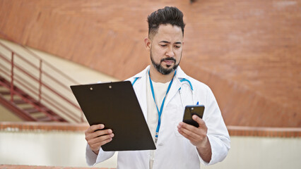 Young latin man doctor using smartphone holding clipboard at hospital