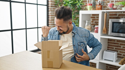 Young latin man unpacking cardboard box with winner expression at dinning room