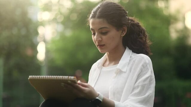Portrait of pretty young indian woman illustrator drawing sketch from nature using brush or pencil outdoors Pretty curly female enjoying beautiful sunny day while painting in green park Art hobby