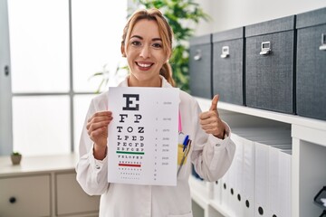 Hispanic optician woman holding medical exam smiling happy and positive, thumb up doing excellent and approval sign