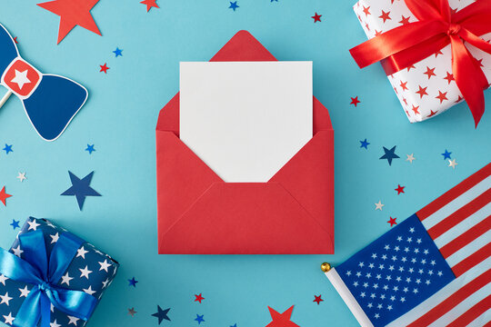 4th of July gift theme concept. Top view flat lay of red envelope with blank card, gift boxes, american flag, party decorations and confetti on pastel blue background with blank space for text