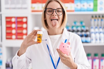 Young caucasian woman working at pharmacy drugstore holding pills an piggy bank sticking tongue out happy with funny expression.