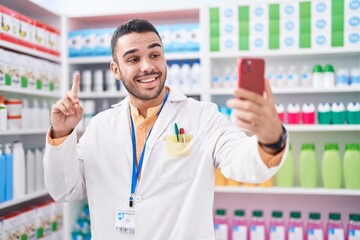 Young hispanic man working at pharmacy drugstore doing video call with smartphone smiling with an idea or question pointing finger with happy face, number one