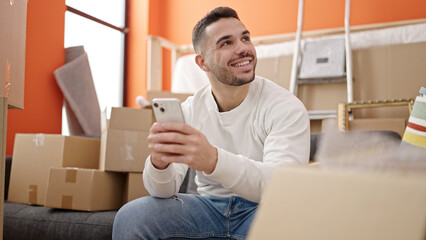 Young hispanic man using smartphone sitting on sofa at new home