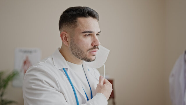 Young hispanic man doctor standing with serious expression taking out medical mask at clinic