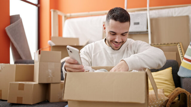 Young hispanic man unpacking cardboard box using smartphone at new home