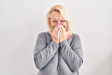 Middle age caucasian woman standing over white background laughing and embarrassed giggle covering...
