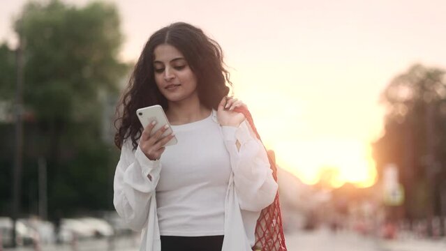 Charming young indian woman with eco bag walking down the street after supermarket and hold smartphone chatting outdoors Happy relaxed lady walking on the city centre with beautiful sunset alone