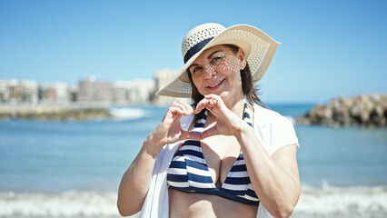 Middle age hispanic woman tourist wearing bikini doing heart gesture with hands at the beach