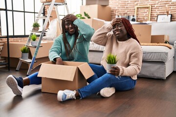 Two african woman moving to a new home sitting on the floor stressed and frustrated with hand on head, surprised and angry face