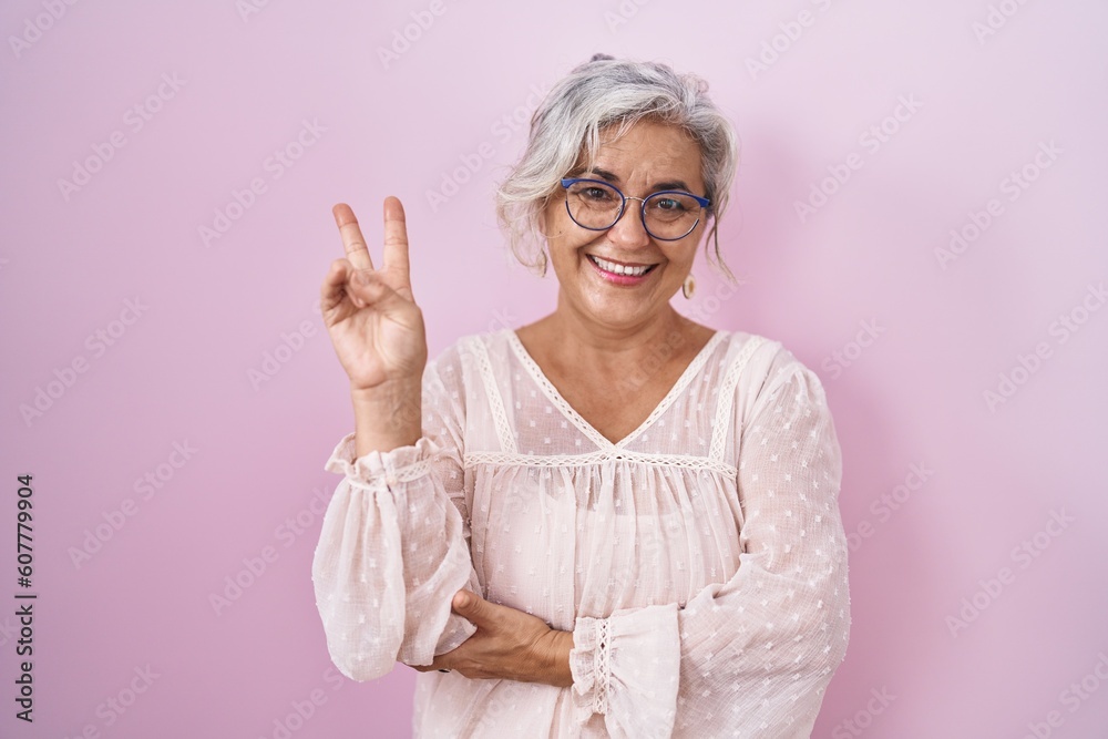 Wall mural Middle age woman with grey hair standing over pink background smiling with happy face winking at the camera doing victory sign. number two.