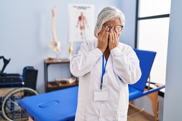 Middle age woman with grey hair working at pain recovery clinic rubbing eyes for fatigue and...