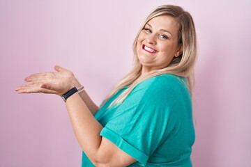 Caucasian plus size woman standing over pink background pointing aside with hands open palms...