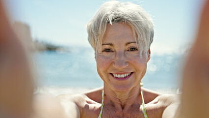 Middle age blonde woman tourist wearing bikini make selfie by camera at the beach
