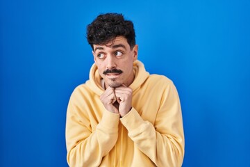 Hispanic man standing over blue background laughing nervous and excited with hands on chin looking to the side