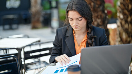 Young beautiful hispanic woman business worker using laptop reading document at coffee shop terrace