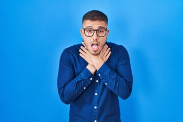 Young hispanic man wearing glasses over blue background shouting and suffocate because painful strangle. health problem. asphyxiate and suicide concept.