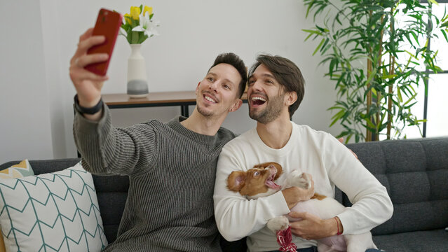 Two men couple making selfie by smartphone sitting on sofa with dog at home