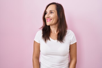 Middle age brunette woman standing over pink background looking away to side with smile on face, natural expression. laughing confident.