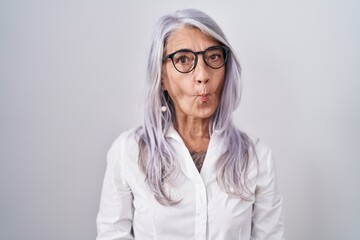 Middle age woman with tattoos wearing glasses standing over white background making fish face with lips, crazy and comical gesture. funny expression.