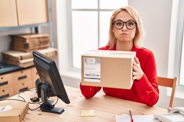 Blonde woman working at small business ecommerce holding box puffing cheeks with funny face. mouth inflated with air, catching air.