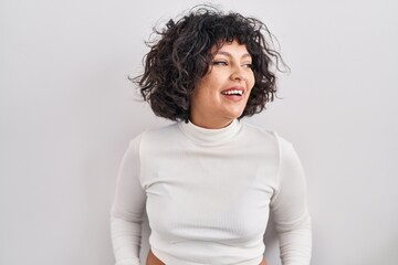 Hispanic woman with curly hair standing over isolated background looking away to side with smile on face, natural expression. laughing confident.