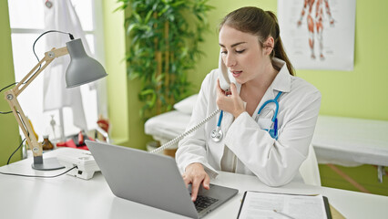 Young beautiful hispanic woman doctor using laptop talking on telephone at clinic