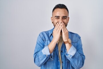 Young hispanic man standing over isolated background laughing and embarrassed giggle covering mouth...