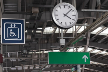 Focus to a blue plate as symbol of lift or elevator for disabled person or wheelchair in train station with blurred background of time on clock and white up arrow on empty green direction light box.