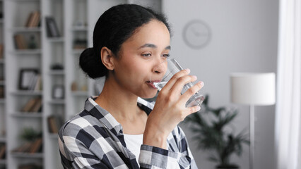 A young African American girl stands by the window in her living room and starts a new day with a good habit, holding a glass of clean water and drinking it, close-up view.