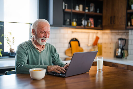 Senior man connected on internet with laptop at home. Senior man with cup of tea using laptop at table in kitchen. Mature man typing on laptop in kitchen during breakfast and driking coffee.