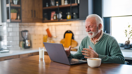Naklejka na ściany i meble Cheerful senior man during video conference in kitchen on the laptop while enjoying breakfast and a cup of coffee. Elderly person using internet online chat technology video webcam making a video call