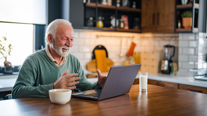 Good-looking enterpreneur working on laptop while having coffee and breakfast. Elderly man using laptop at kitchen table. Computer, money and communication with an elderly male pensioner