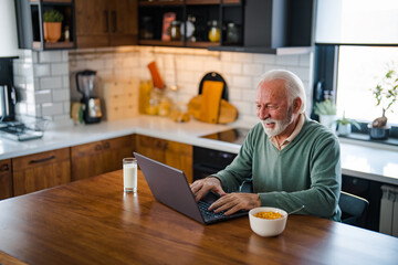 Busy with work. Grey-haired bearded senior man eyeglasses looking concentrated. One senior businessman standing in the kitchen at home and working on the laptop while drinking coffee.