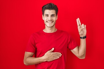 Naklejka premium Young hispanic man standing over red background smiling swearing with hand on chest and fingers up, making a loyalty promise oath