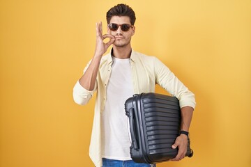 Young hispanic man holding suitcase going on summer vacation mouth and lips shut as zip with...