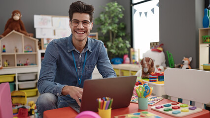 Young hispanic man preschool teacher using laptop sitting on table at kindergarten